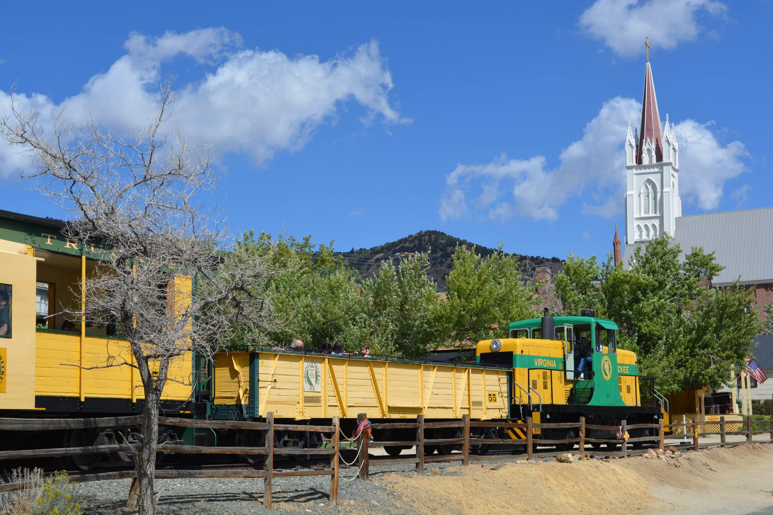 Yellow freight cars and a green locomotive sit in a rail yard, with a white church steeple against a blue sky in the background.