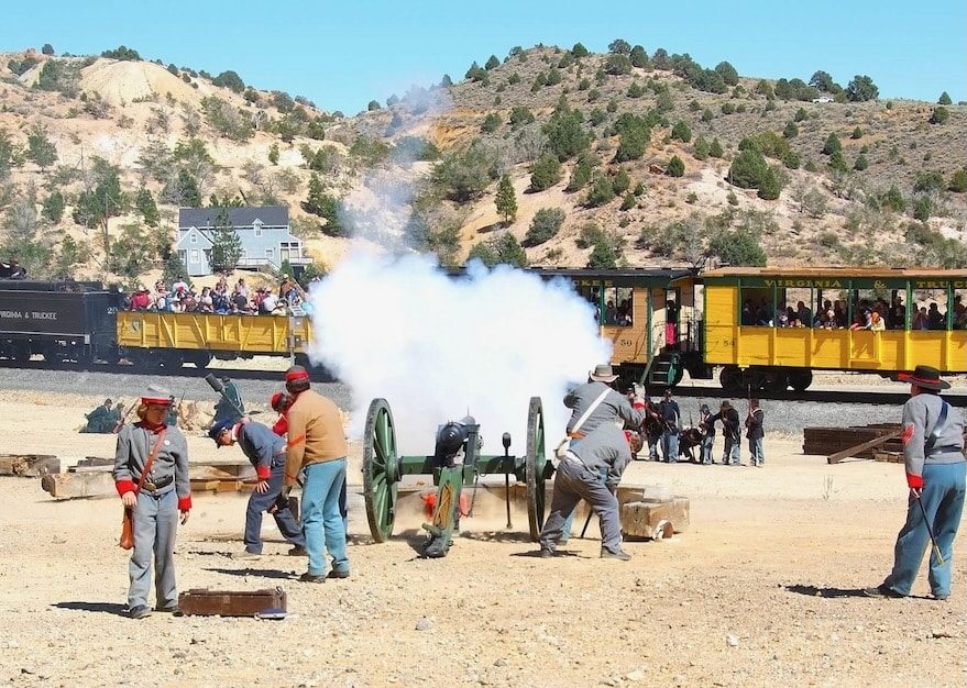 battle2 1 | Virginia & Truckee Railroad A historical reenactment: soldiers fire a large black cannon as smoke billows, with a yellow passenger train and spectators in the background.