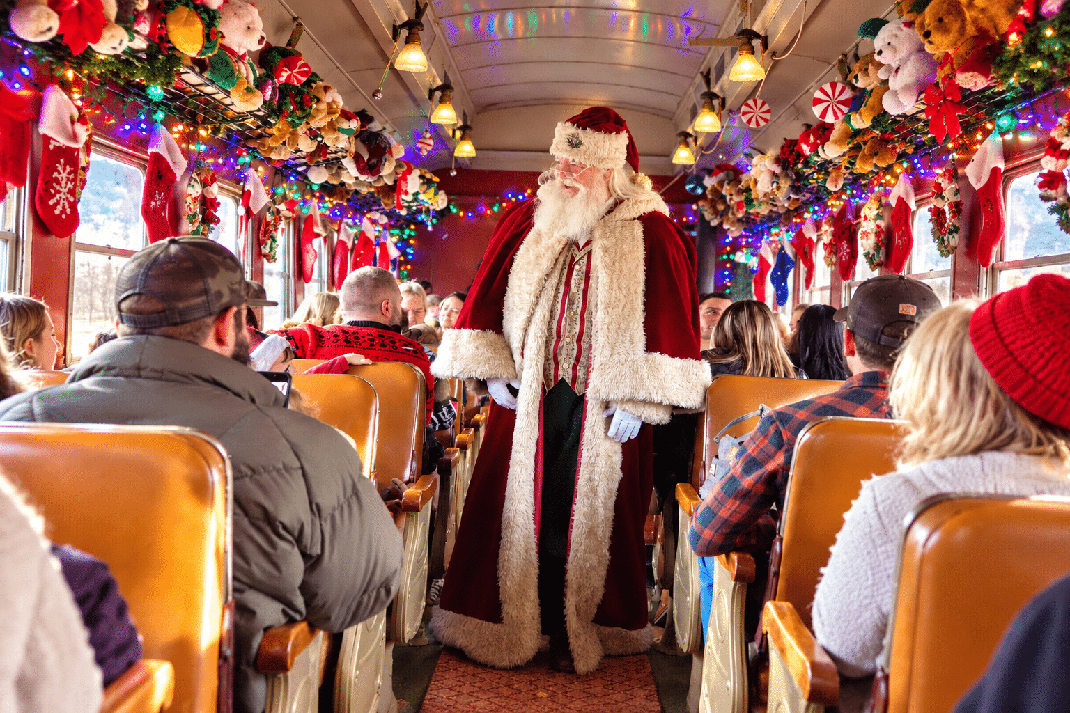 Santa Claus in a red fur-trimmed suit stands in a festively decorated bus aisle among seated passengers. (Informative)